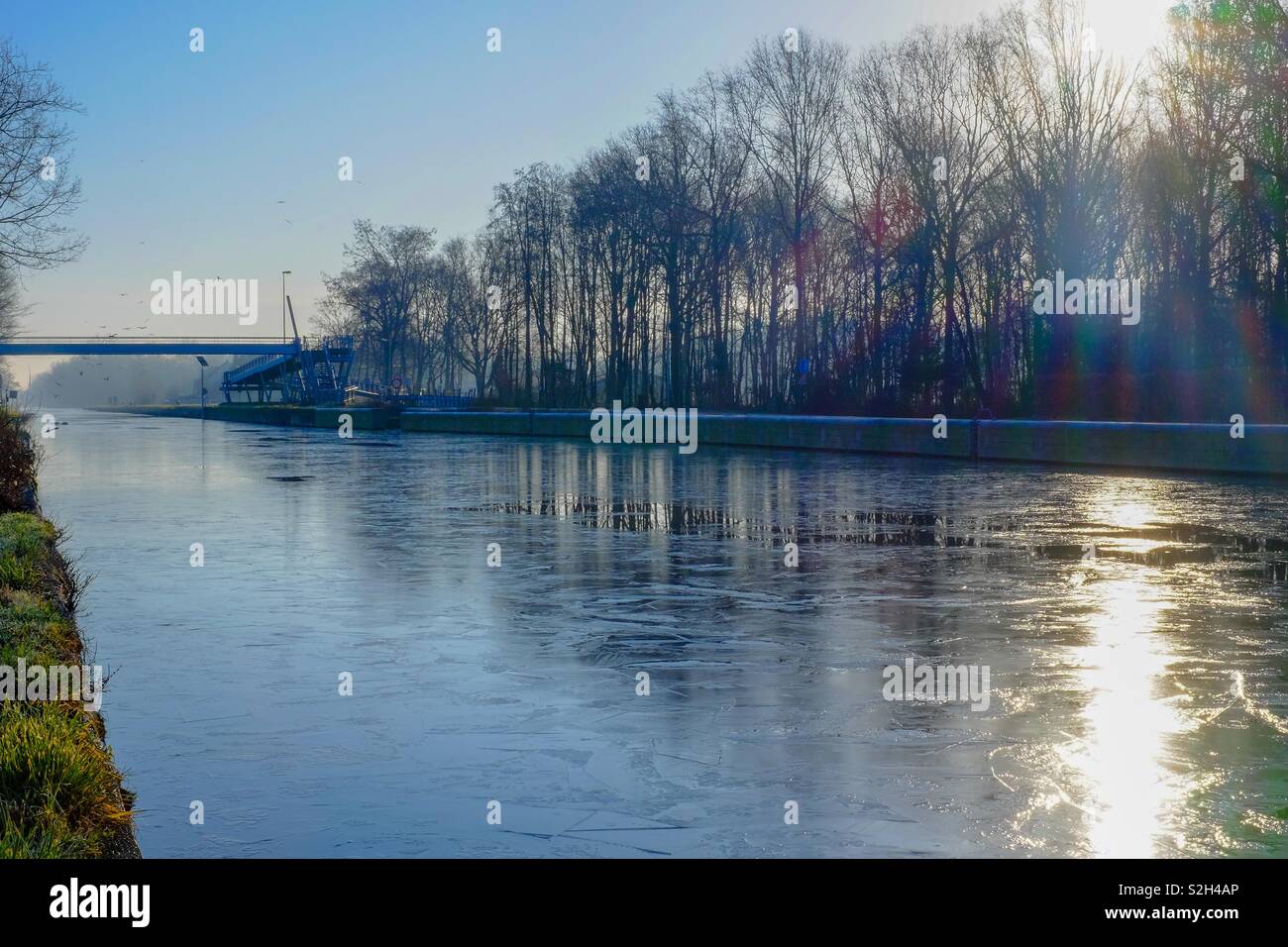 Winter sunrise over a frozen river running between the forest with the sun and the blue sky reflected on the ice and water - Smartphone Captured Stock Image