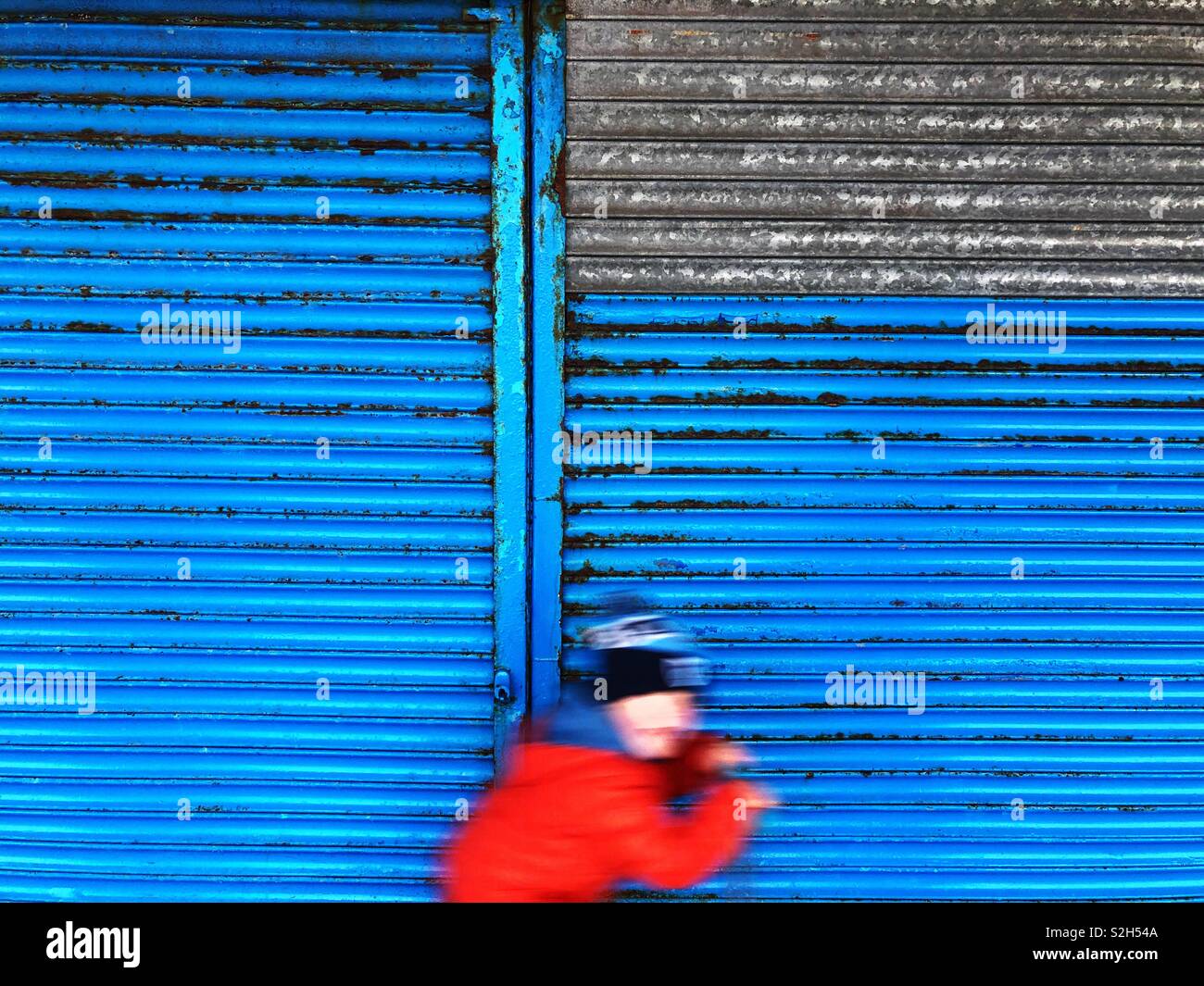 Boy on a scooter passing by a blue roller shutter. - Smartphone Captured Stock Image