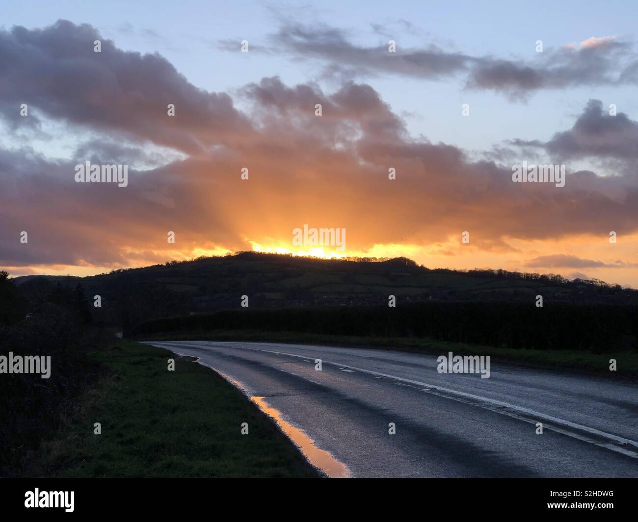 Dramatic sunset behind Winchcombe lighting up low cloud and the hill horizon with intense orange colours and crepuscular rays as well as being reflected in a puddle on the road. - Smartphone Captured Stock Image