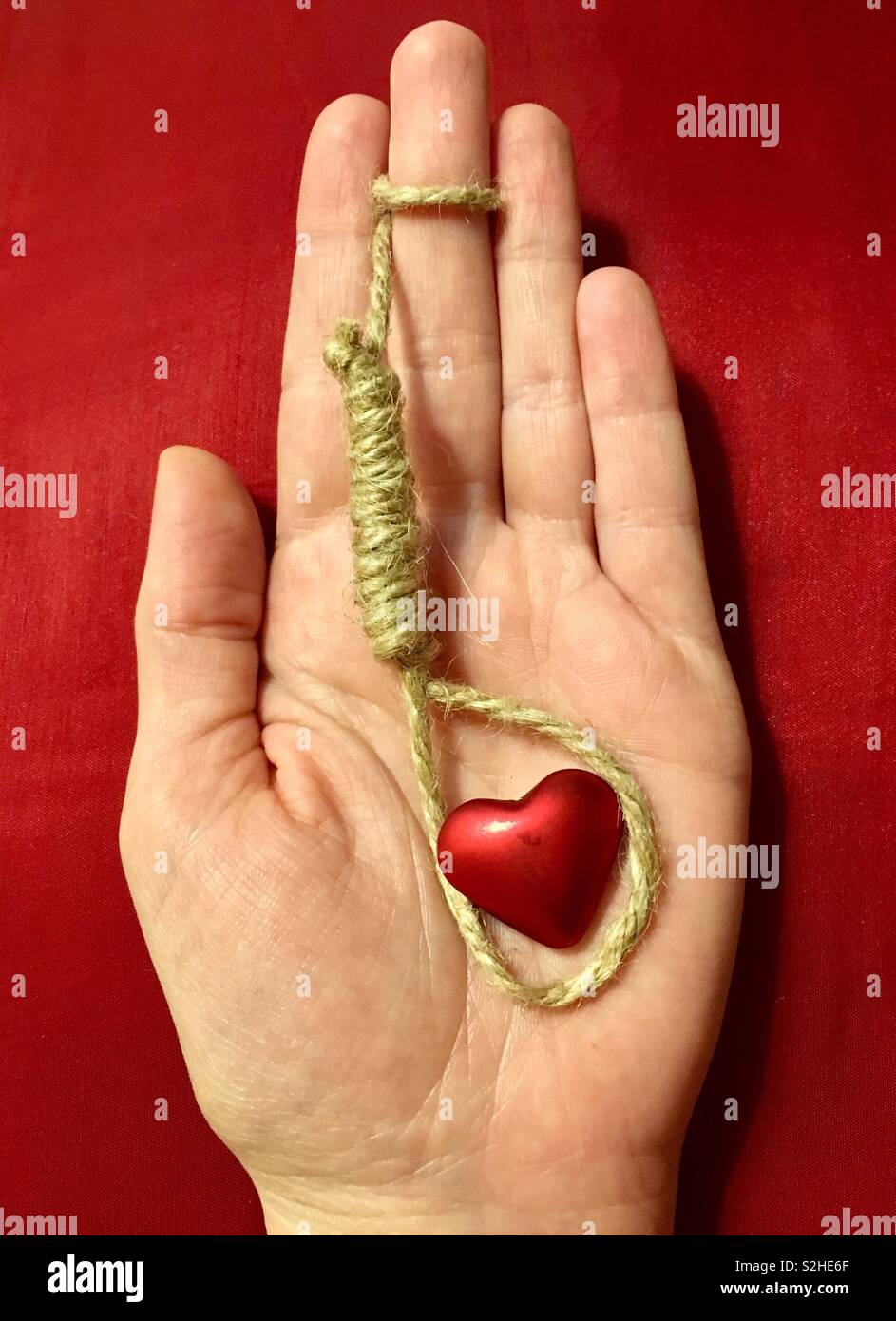 You’re suffocating me: hand holding hangman’s noose around red heart on red background, concept of fatal attraction Stock Photo