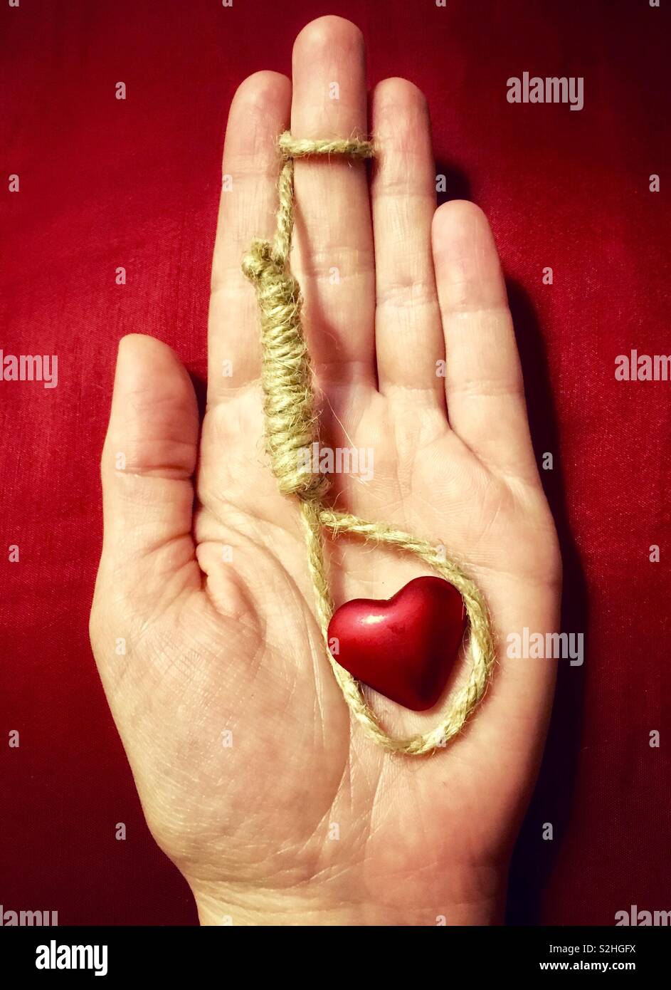 You’re suffocating me: hand holding hangman’s noose around red heart on red background, concept of fatal attraction - Smartphone Captured Stock Image