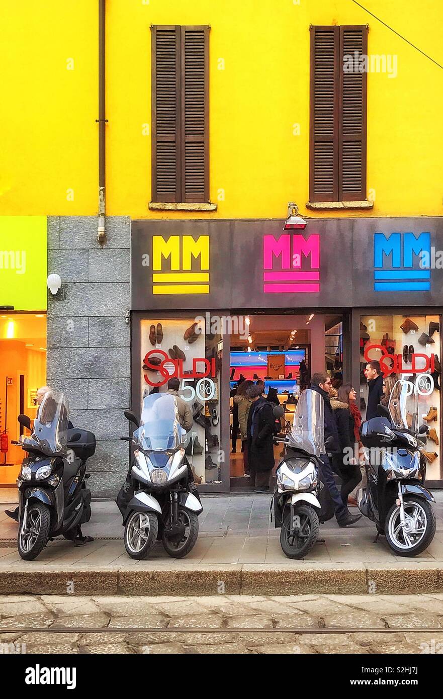 Motorcycles parked on the pavement in front of a shoe and accessories shop MM in central Milan on a busy Saturday morning in winter - Smartphone Captured Stock Image