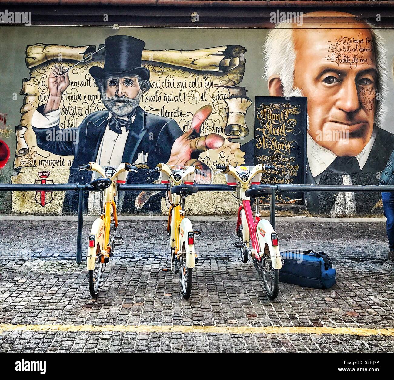 Three bicycles parked in a cycle rack on the cobbled sidewalk in front of mural art on side of a building in Milan, Italy. A bag is on the sidewalk next to one of the bikes, belonging to a young man Stock Photo