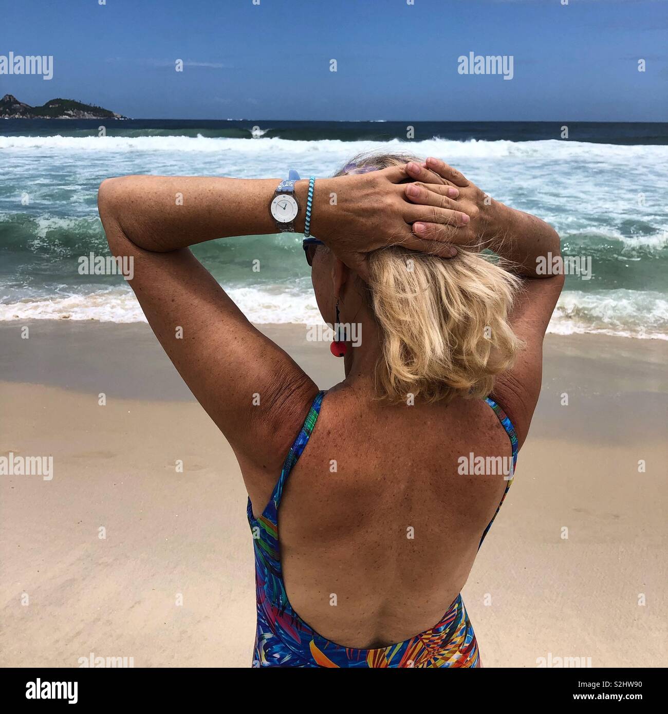 Woman with blond hair on beach looking towards the sea, hands behind her head. - Smartphone Captured Stock Image