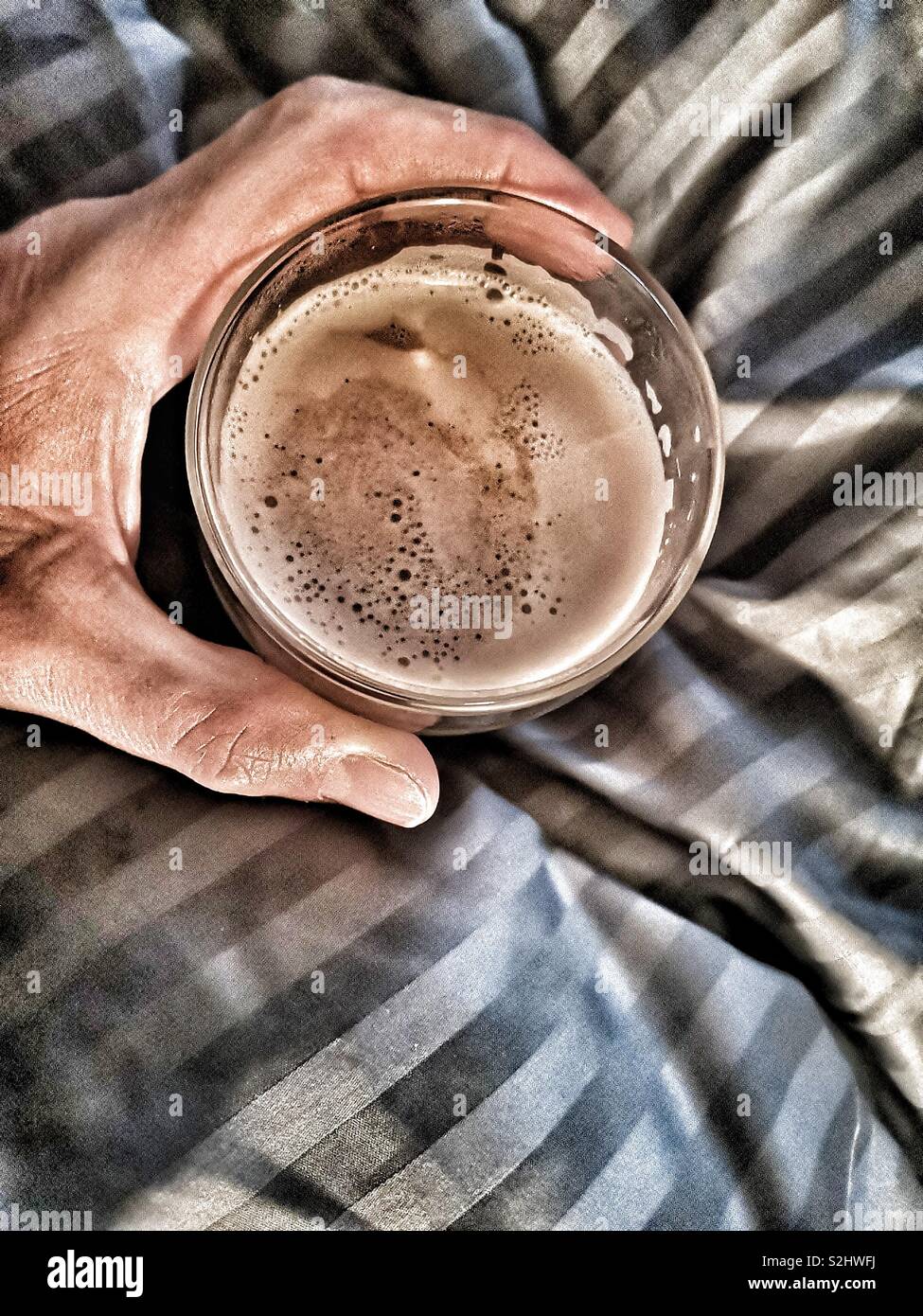 Top view of man’s hand holding glass of cafe latte in bed - Smartphone Captured Stock Image