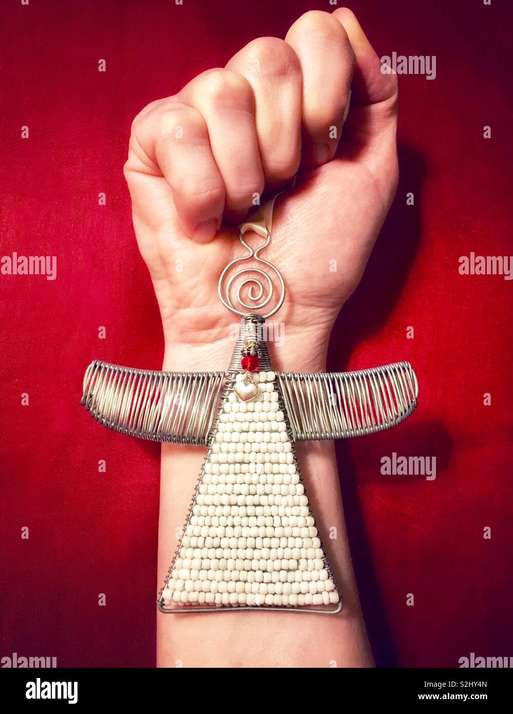 Woman’s hand holding handmade wire and beaded angel ornament with red heart against red background - Smartphone Captured Stock Image