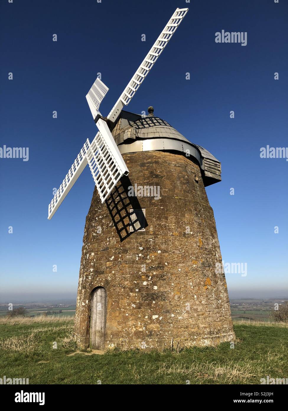 A stone windmill with white sails at the top of a lofty hill with expansive views on the northern edge of the Cotswolds framed by a brilliant blue sky on a Spring day. - Smartphone Captured Stock Image