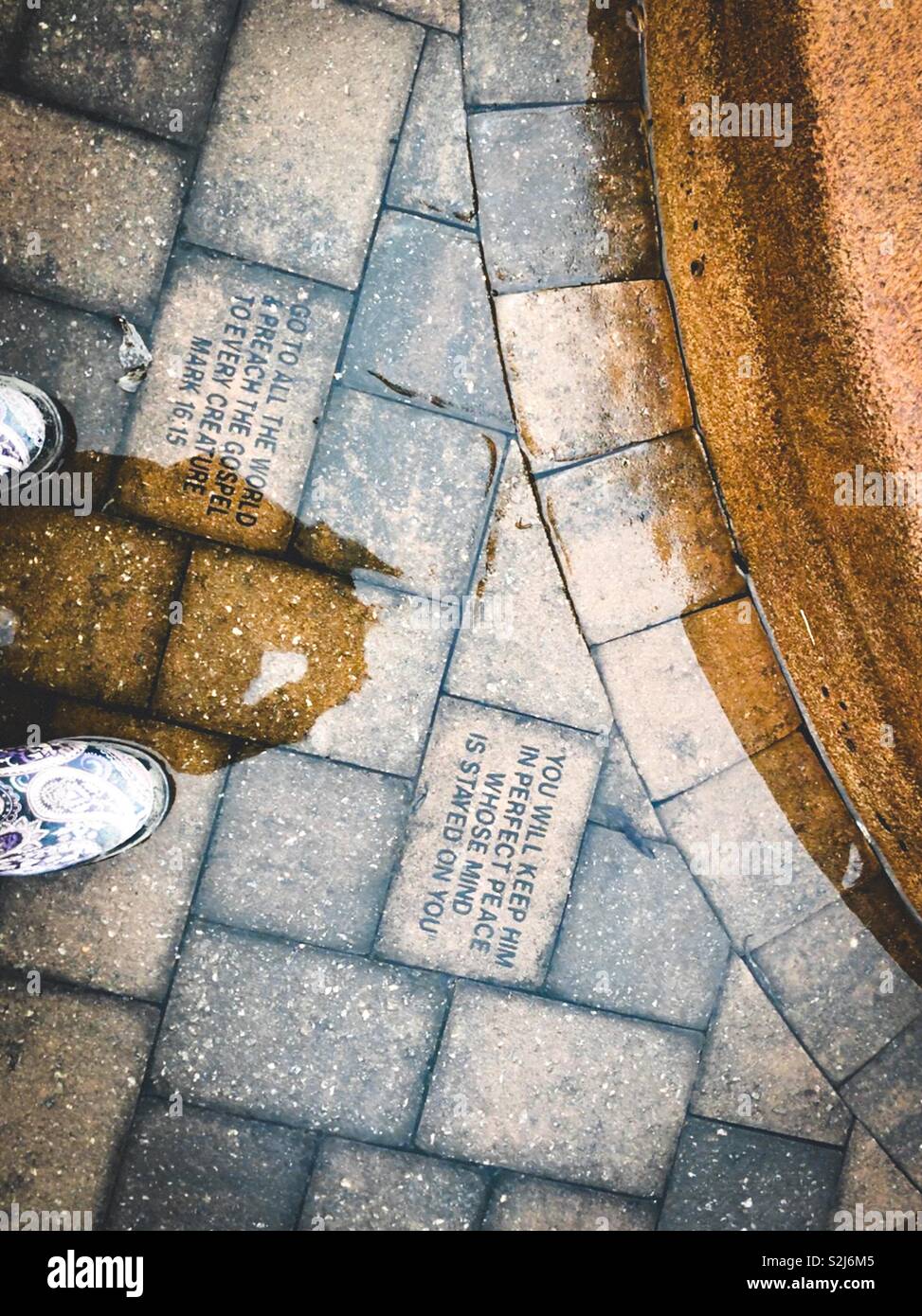 Shadows and boots in photo of scripture verses etched in pavers beside fountain base on a rainy day - Smartphone Captured Stock Image