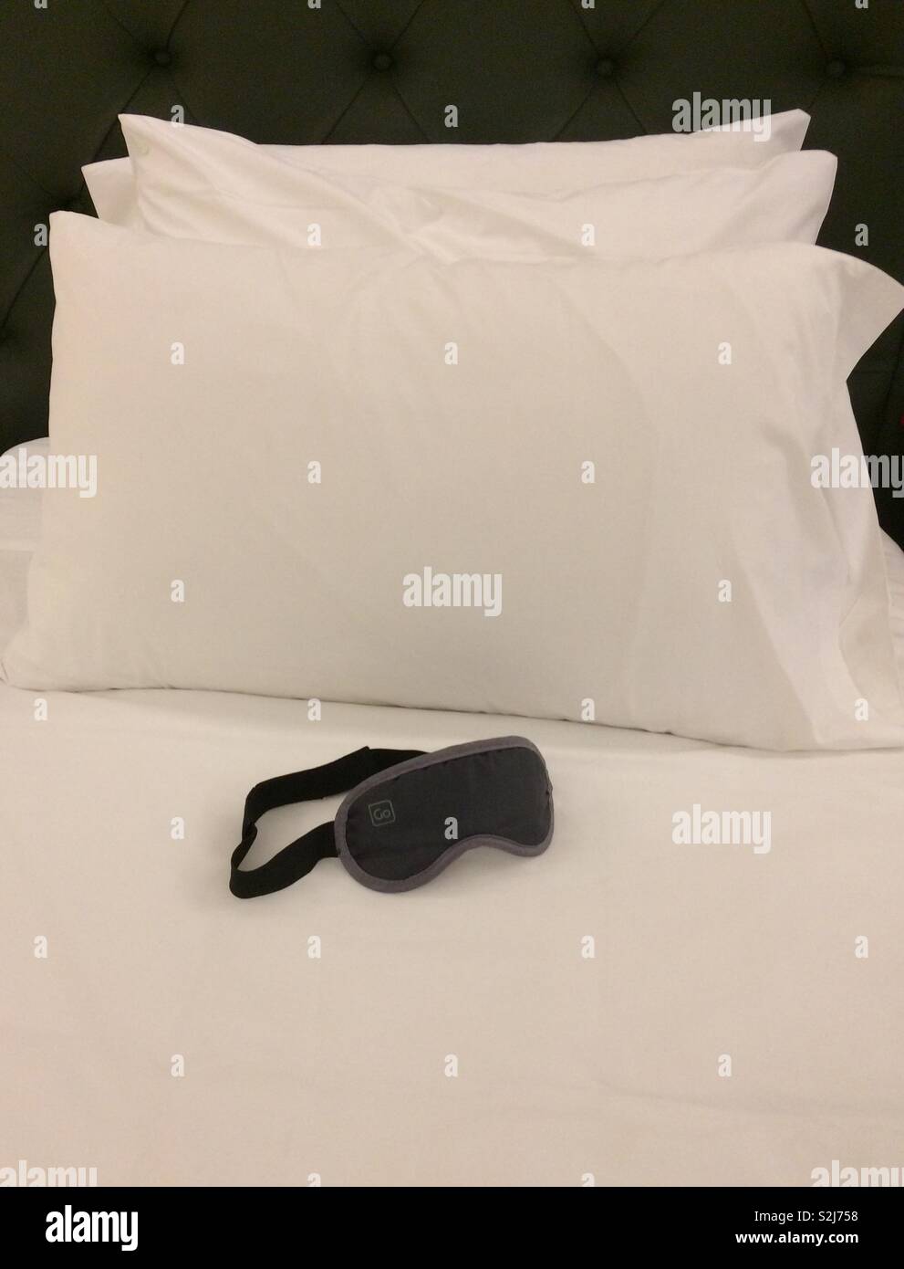 Clean, fresh, white hotel bed linen and three soft pillows on a single bed with eyeshades ready - a study in black and white perfection, and awaiting a sleeper. Stock Photo