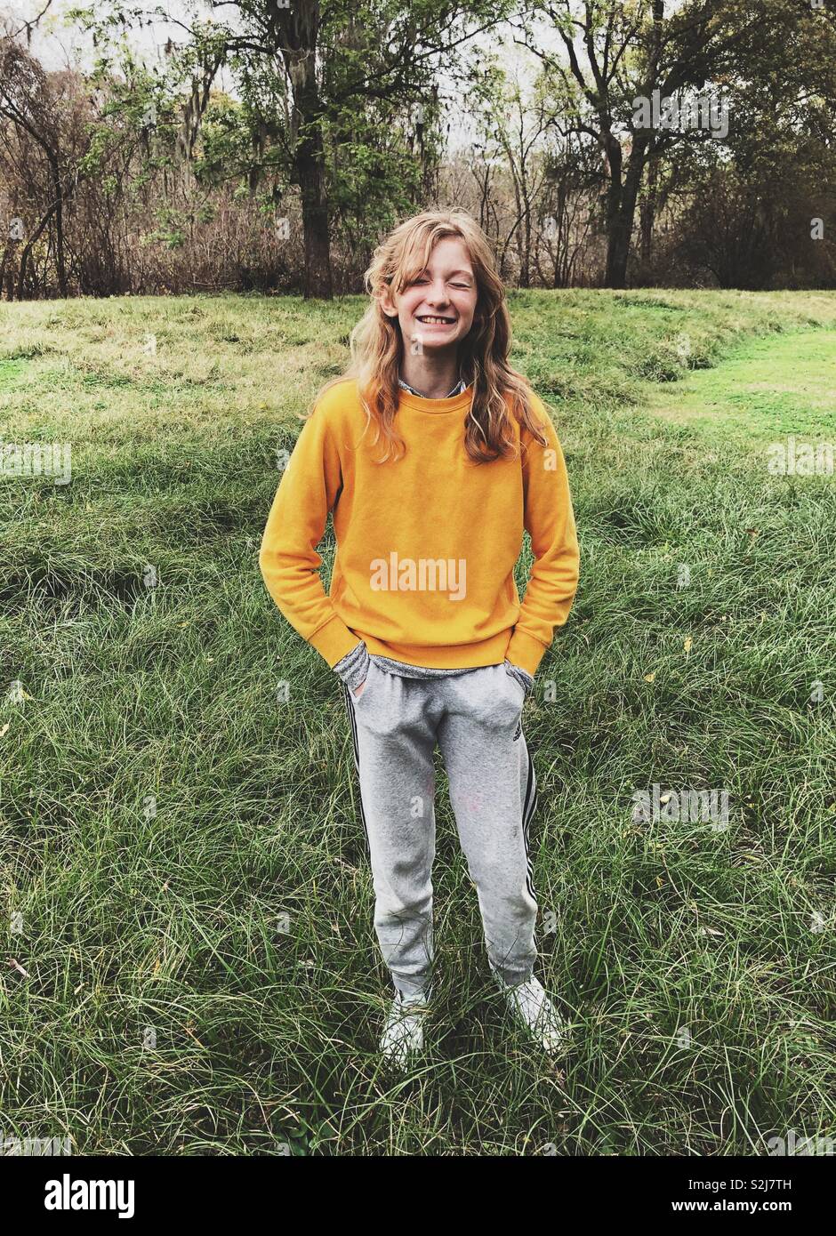 Young blond girl wearing yellow sweatshirt, smiling and squinting her eyes in a meadow by the forest Stock Photo