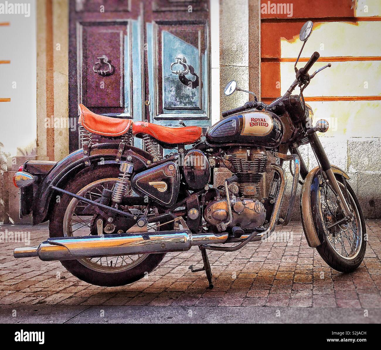 A vintage Royal Enfield motorbike parked on a cobblestone pavement in front of the entrance of a building. The entrance comprises a wooden door Stock Photo