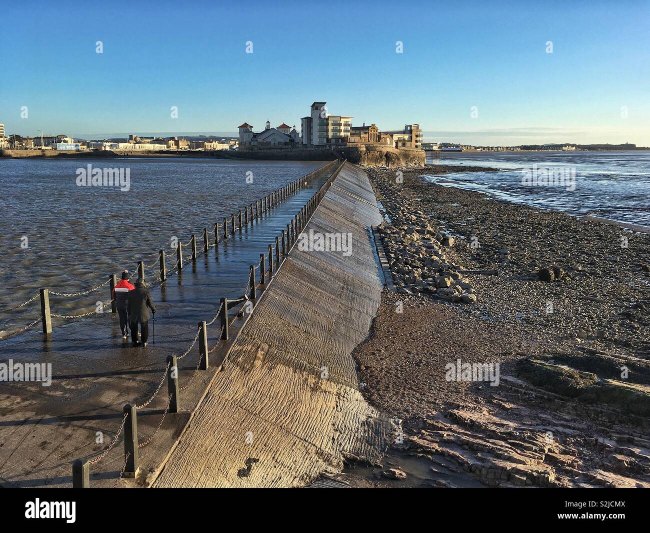 Two walkers cross the Marine Lake causeway in Weston-super-Mare, UK on a sunny winter afternoon. - Smartphone Captured Stock Image