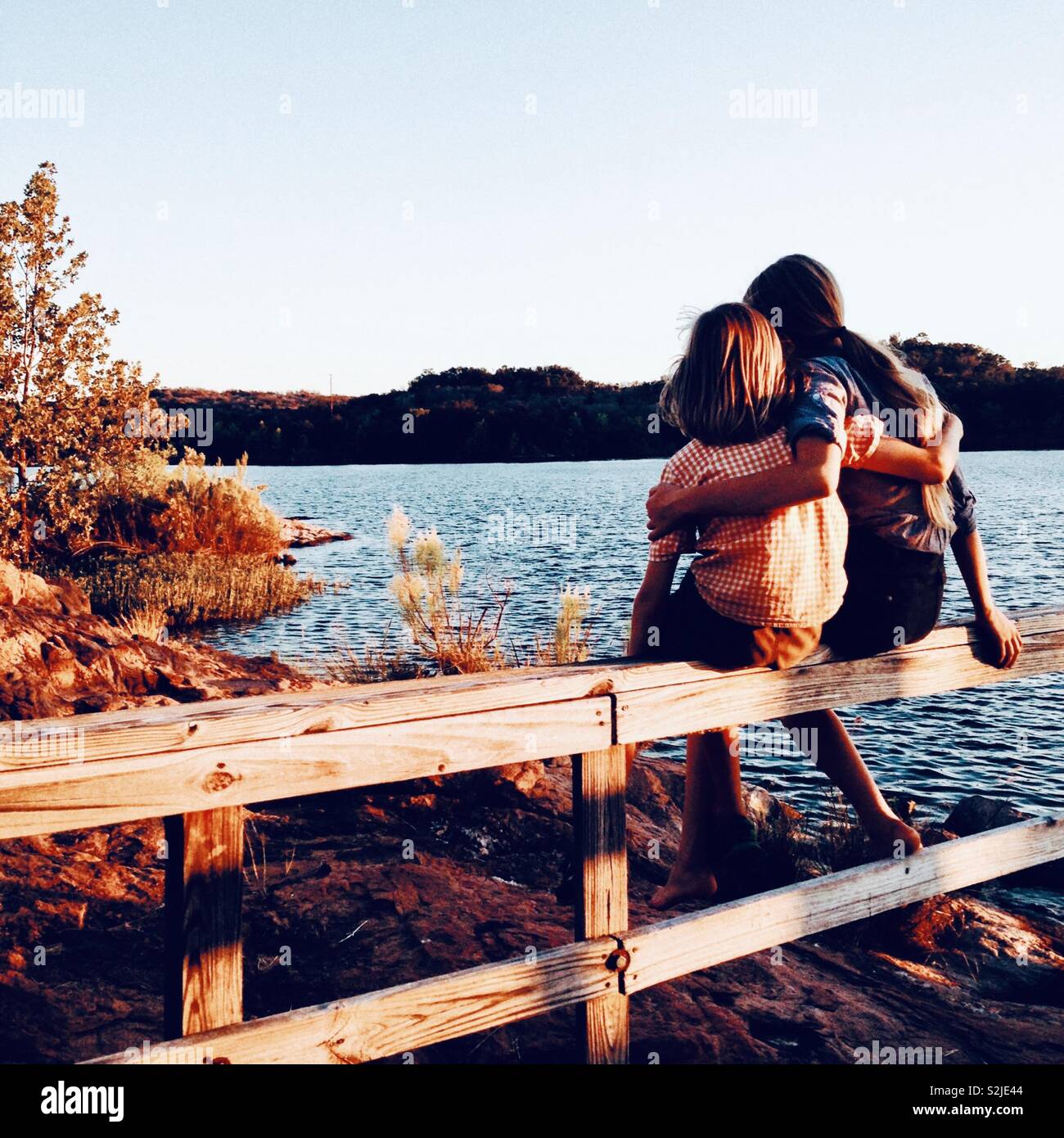 Boy and a girl, brother and sister sitting close on a wood rail by the lake at sunset Stock
