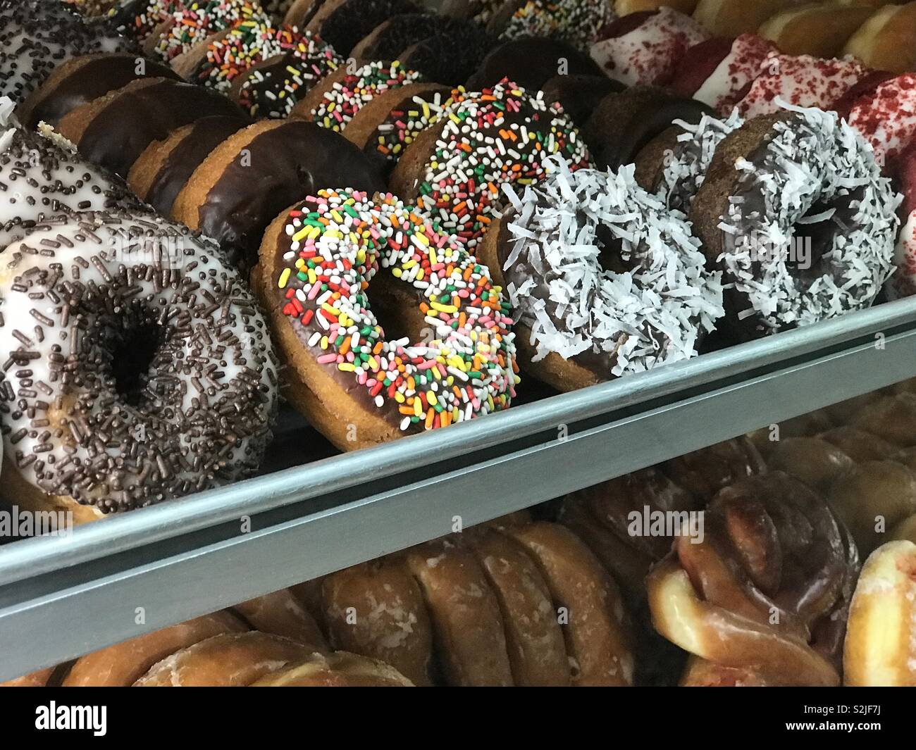 A variety of fresh cake donuts with various toppings (sprinkles, coconut, red velvet) are shown in a bakery display case. - Smartphone Captured Stock Image