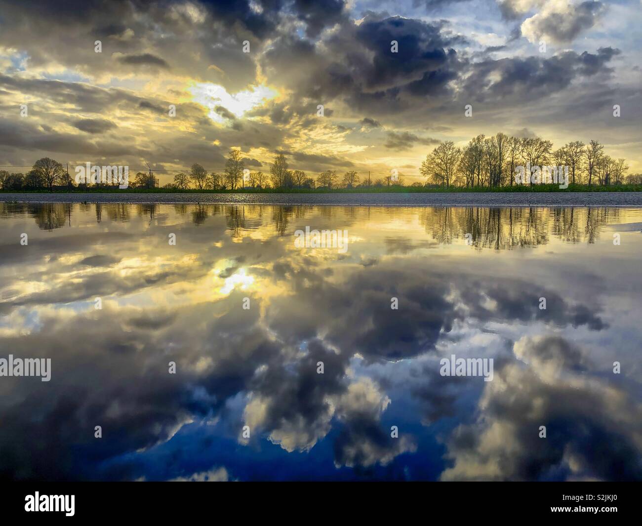 Colorful and dramatic sunset in a cloudy sky with white and dark clouds reflected on the surface of the water of a puddle after the rain - Smartphone Captured Stock Image