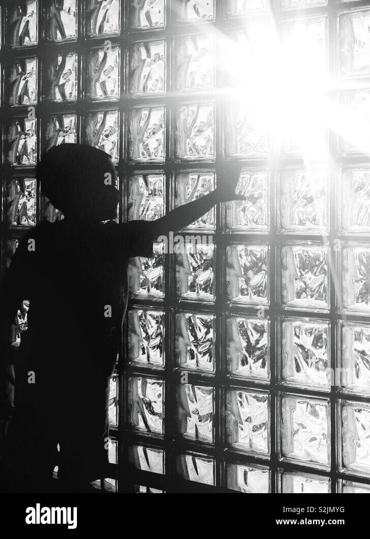 Silhouette of a boy reaching out to touch the sunlight shining through a glass window wall.  Black and white.  Inspiration investigating. - Smartphone Captured Stock Image