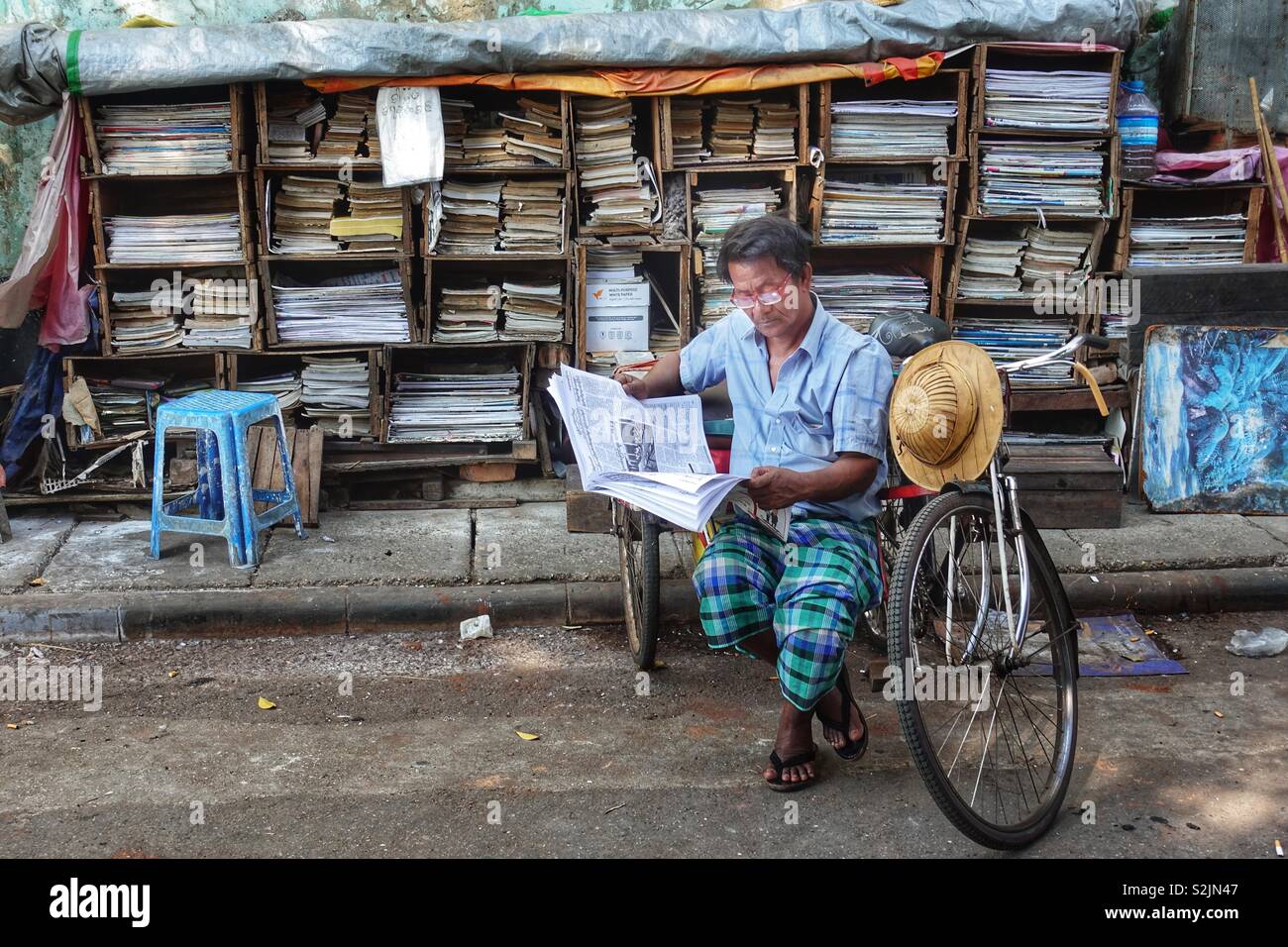 Myanmar everyday life: portrait of an adult man reading the newspaper. Sitting next to a book stand and in his bicycle Rikscha. On the streets of Yangon, Burma. Stock Photo