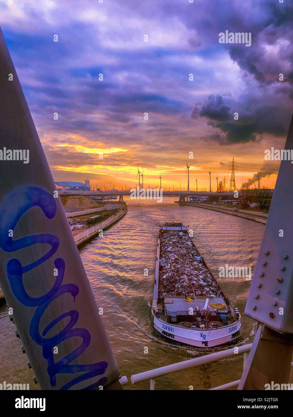 Large river transport boat going underneath a bridge over the river scheldt at sunset showing the Golden glow in the sky under the dark and blue sky above - Smartphone Captured Stock Image