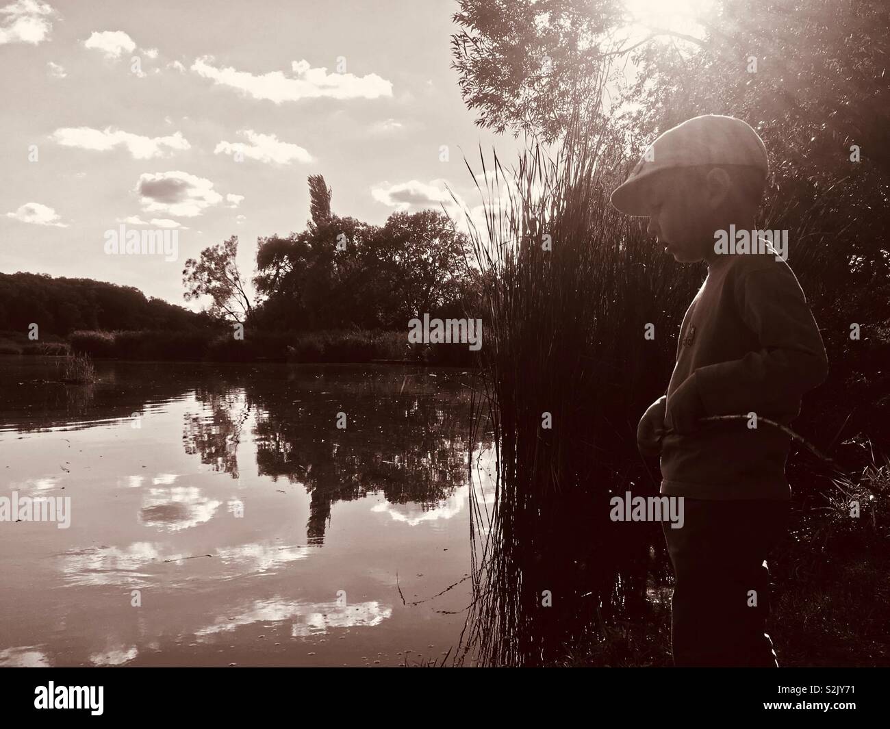 Boy child standing on the bank of a small fishing lake at sunset - Smartphone Captured Stock Image
