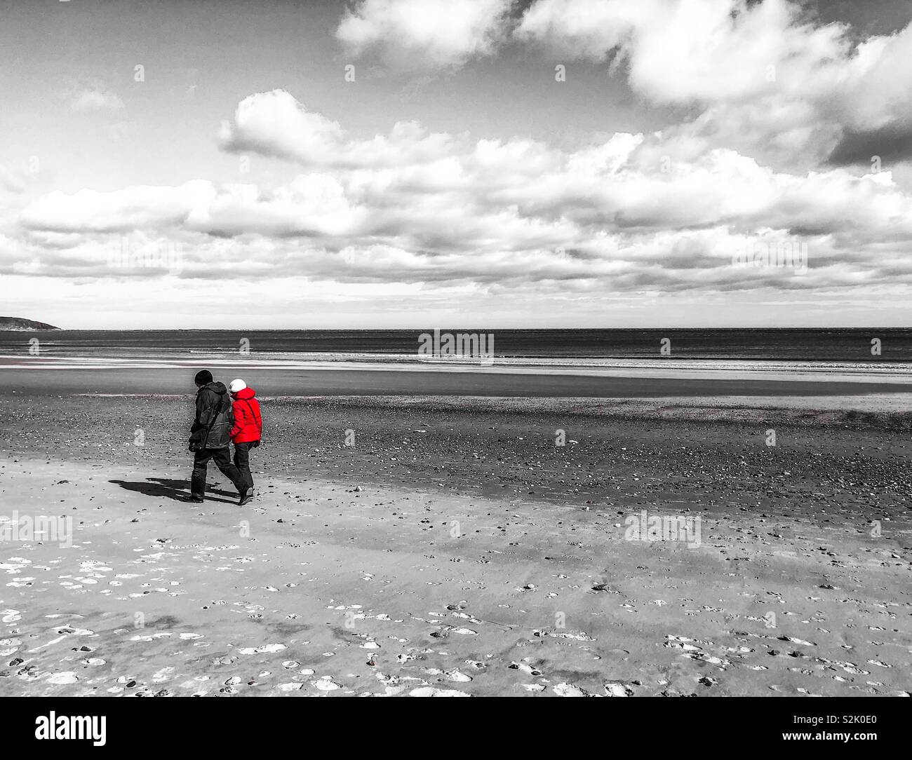 Couple walking along a beach in winter. Black and white image with a pop of red on the woman’s coat - Smartphone Captured Stock Image