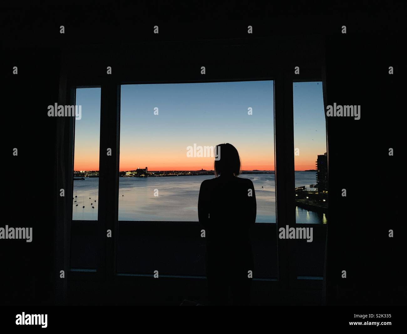 A young women looks out a window at Boston Harbor at sunrise. Boston, Massachusetts USA. Stock Photo