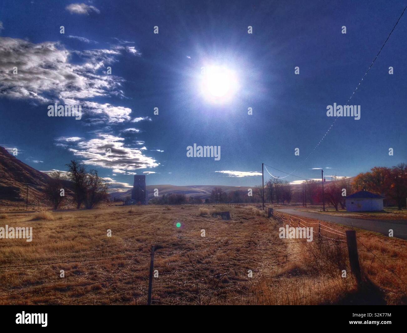 Deep blue sky and midday sun over dirt road and distant grain elevator and hillside in Eastern Washington - Smartphone Captured Stock Image