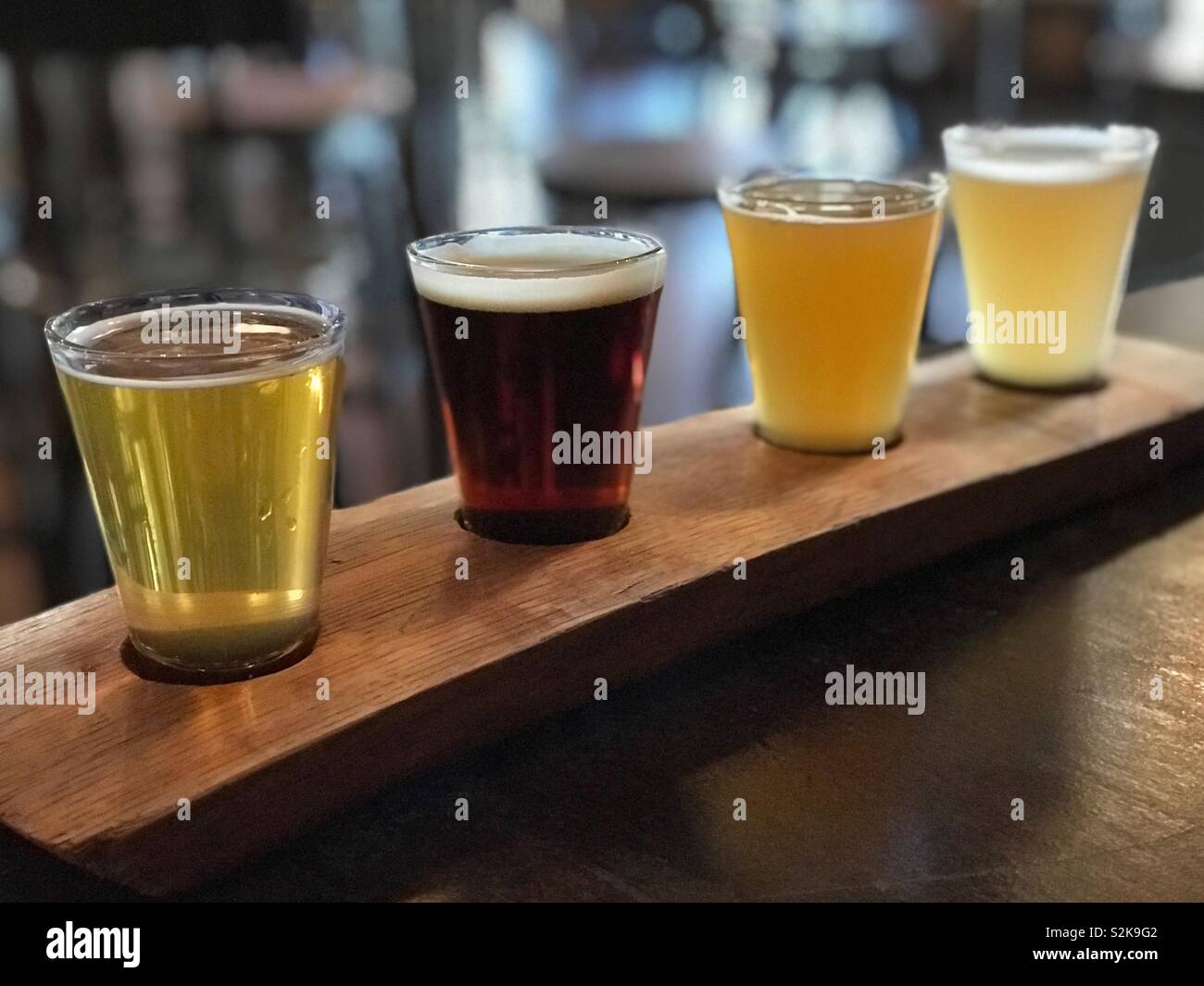 A sampler of four small glasses filled with craft beer is shown on a wood flight paddle in a restaurant. - Smartphone Captured Stock Image