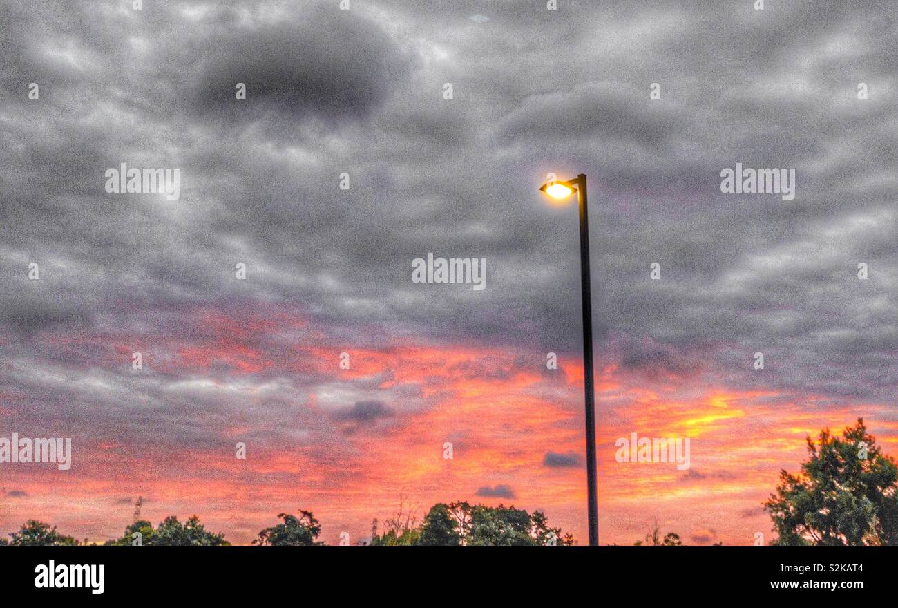 Streetlight lit up during the orange glow of a sunset with grey clouds Stock Photo