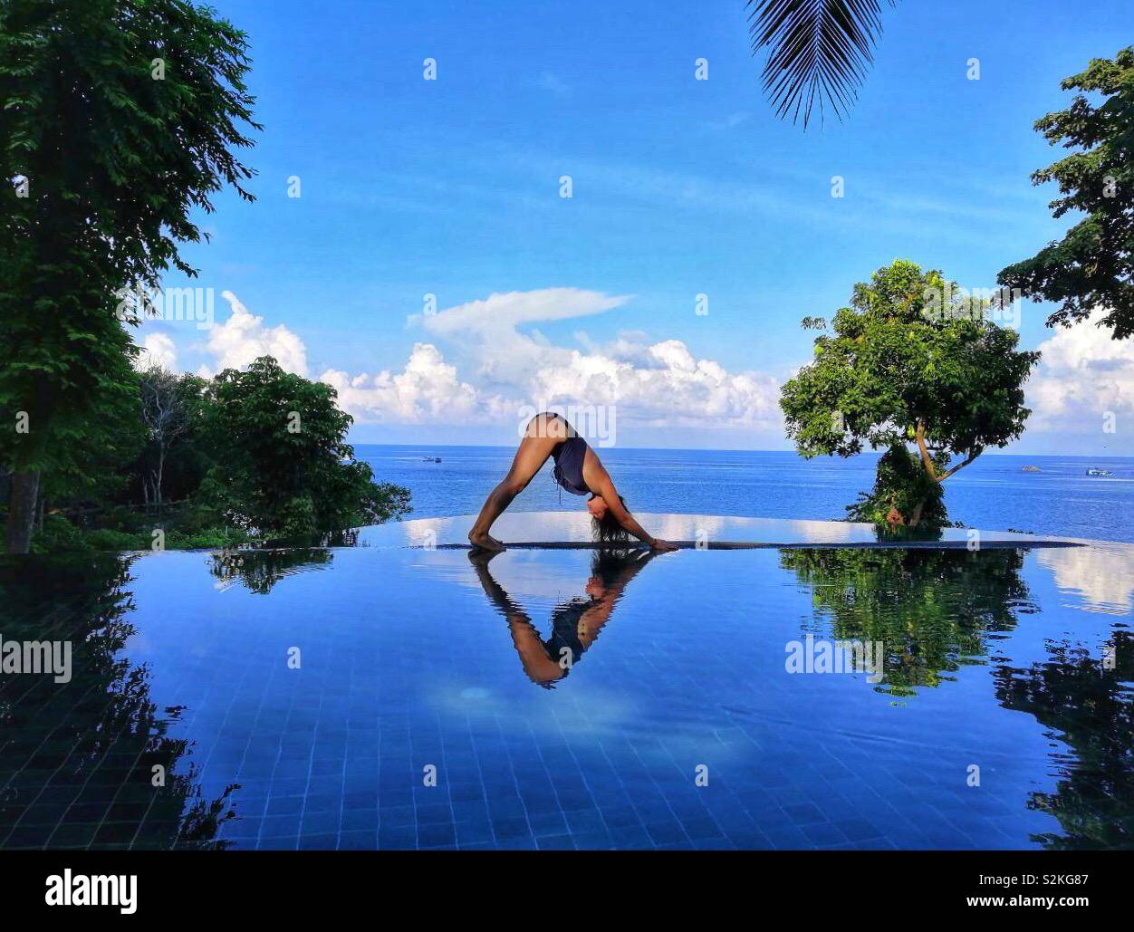 Woman making yoga dog pose by the swimming pool with sea background, Thailand - Smartphone Captured Stock Image