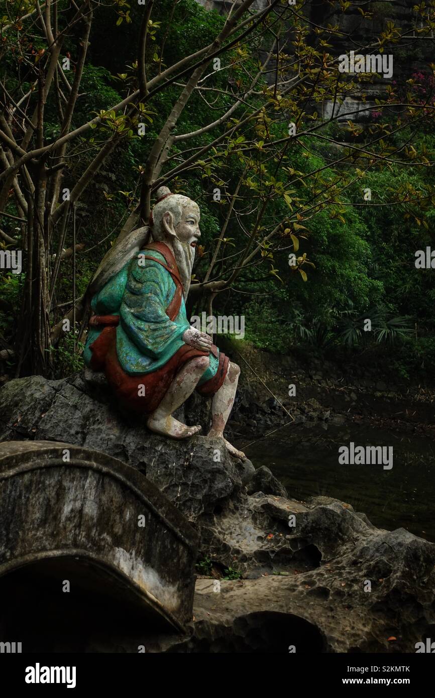 Artful asian still life: statue of an old and bearded Vietnamese monk. Or traveler. Resting on a small bridge and looking to the horizon. Stock Photo
