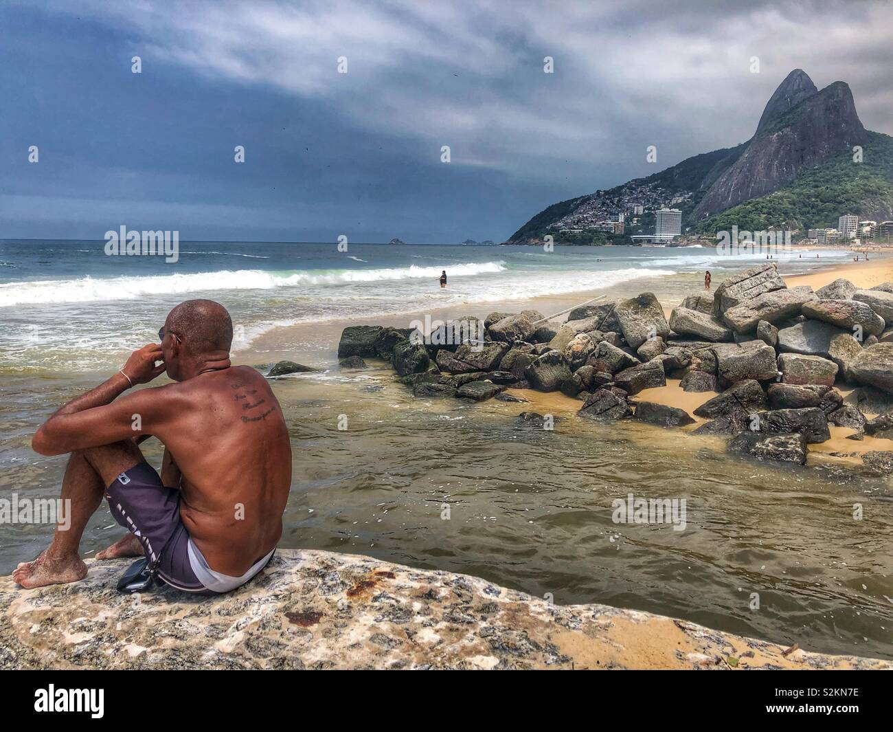 A man sits on n the rocks that divide Ipanema and LeBlon beaches in Rio de Janeiro, Brazil. - Smartphone Captured Stock Image