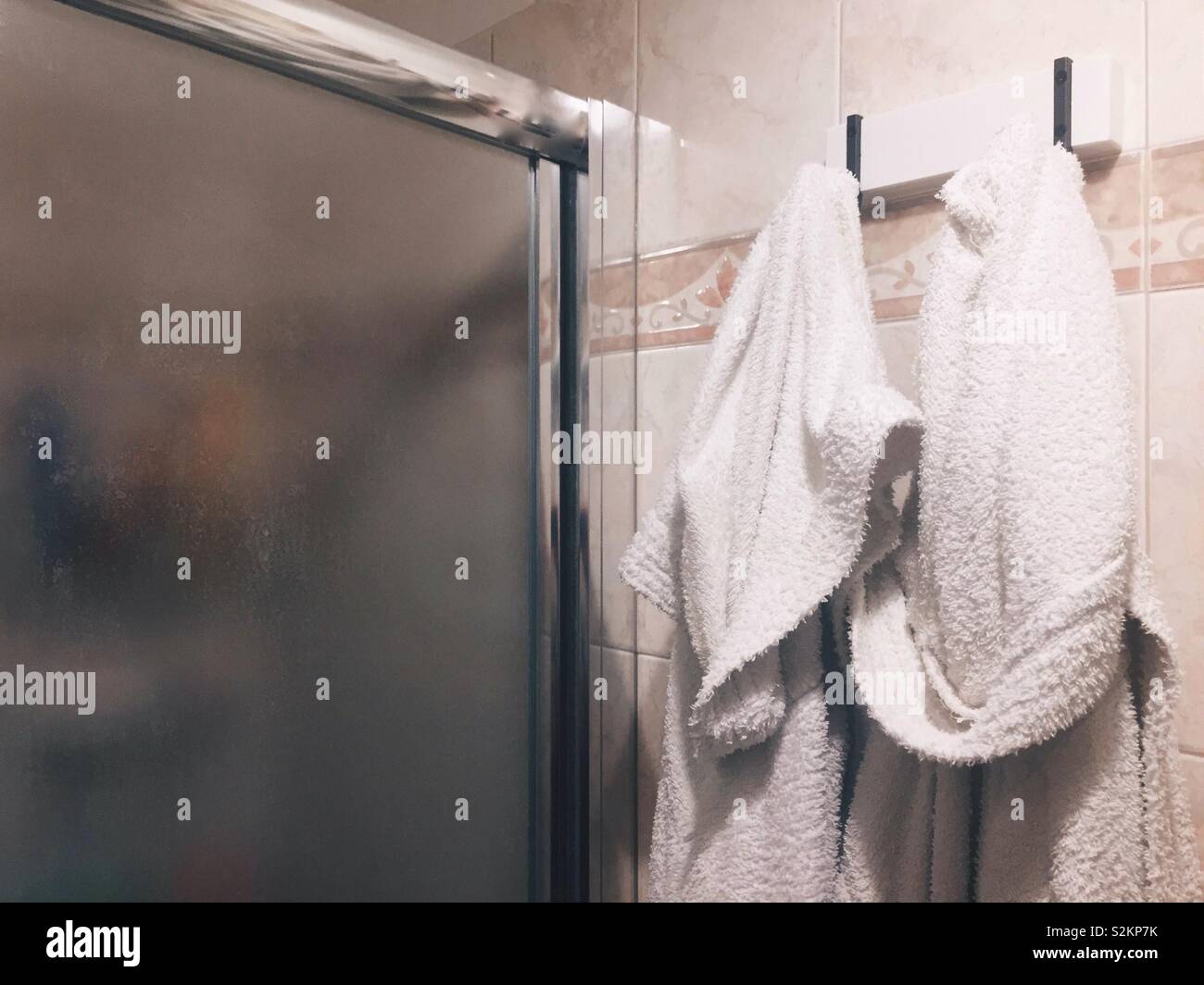 A couple of white bathrobes hanging on the wall near the glass door of a shower stall Stock Photo