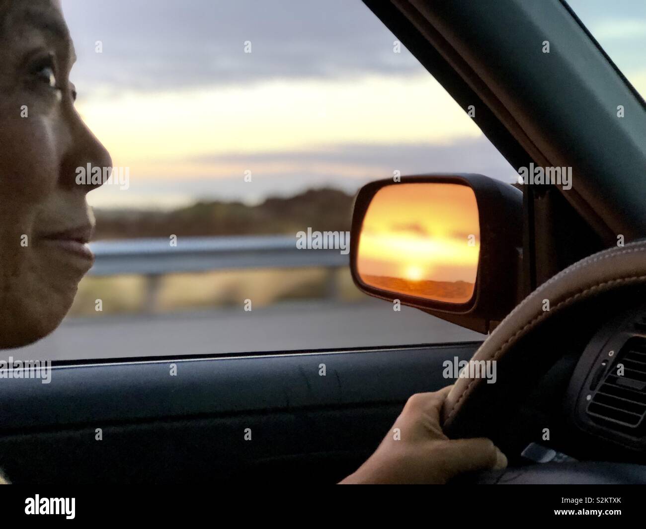Asian woman driving while sun is setting in rear view mirror. - Smartphone Captured Stock Image