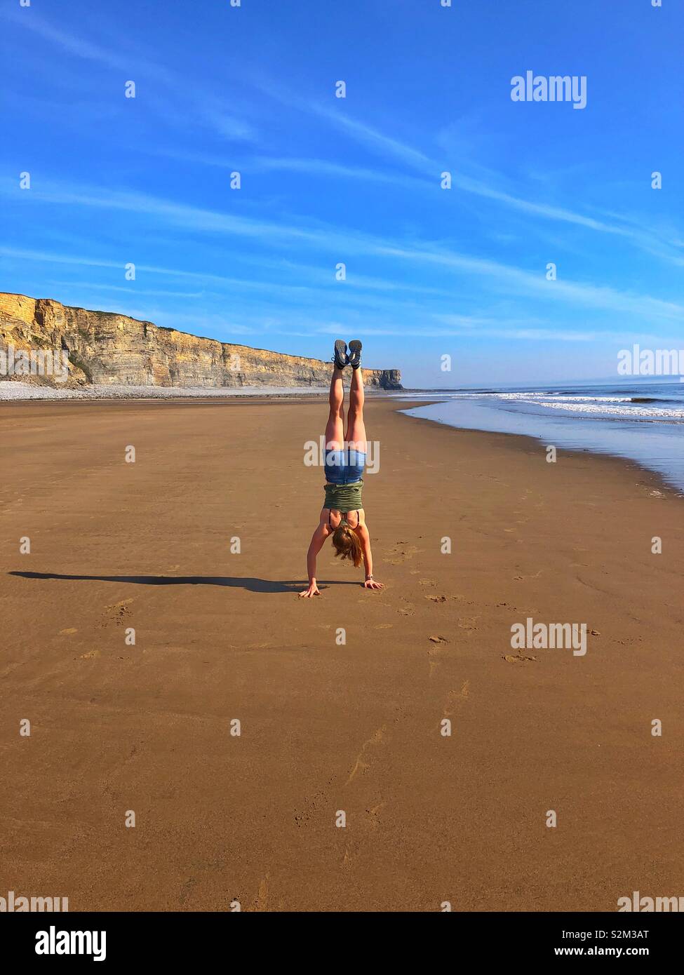 Young woman doing a handstand on a large sandy beach, South Wales, April. - Smartphone Captured Stock Image