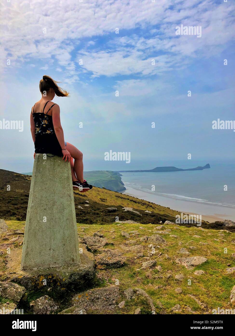Young woman sitting on the trig point of Rhossili downs enjoying the view above Rhossili Bay, Gower, Swansea, Wales. - Smartphone Captured Stock Image