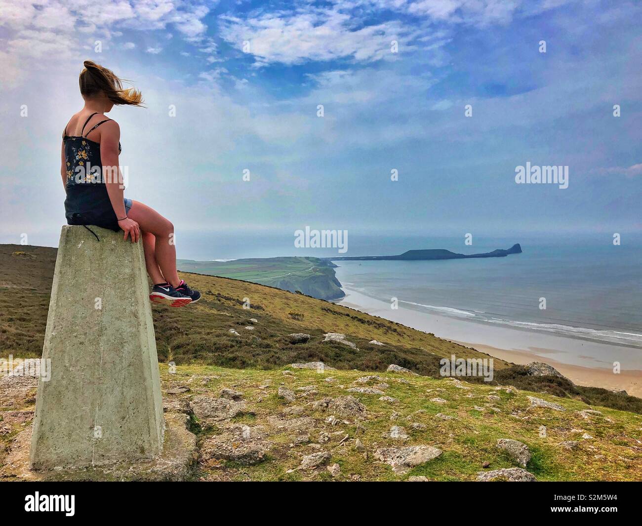 Young woman sitting on the trig point on Rhossili downs enjoying the view over Rhossili Bay, Gower, Wales, April. - Smartphone Captured Stock Image
