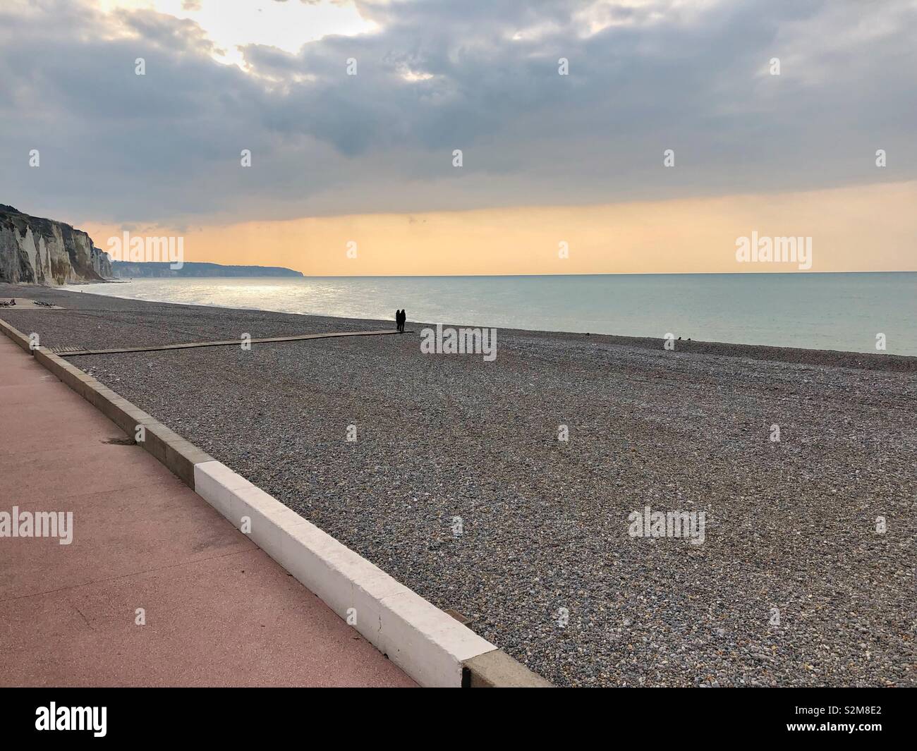 A couple at the end of a wooden walkway on The otherwise empty beach on a chilly April evening in Dieppe, France. - Smartphone Captured Stock Image