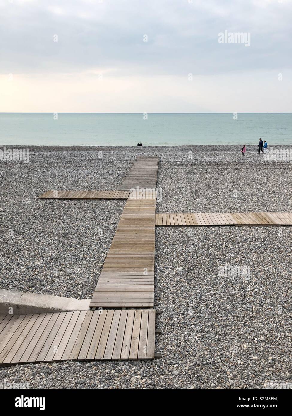 A few people walking on the pebble beach in Dieppe in off season with a wooden walkway in the foreground. Stock Photo