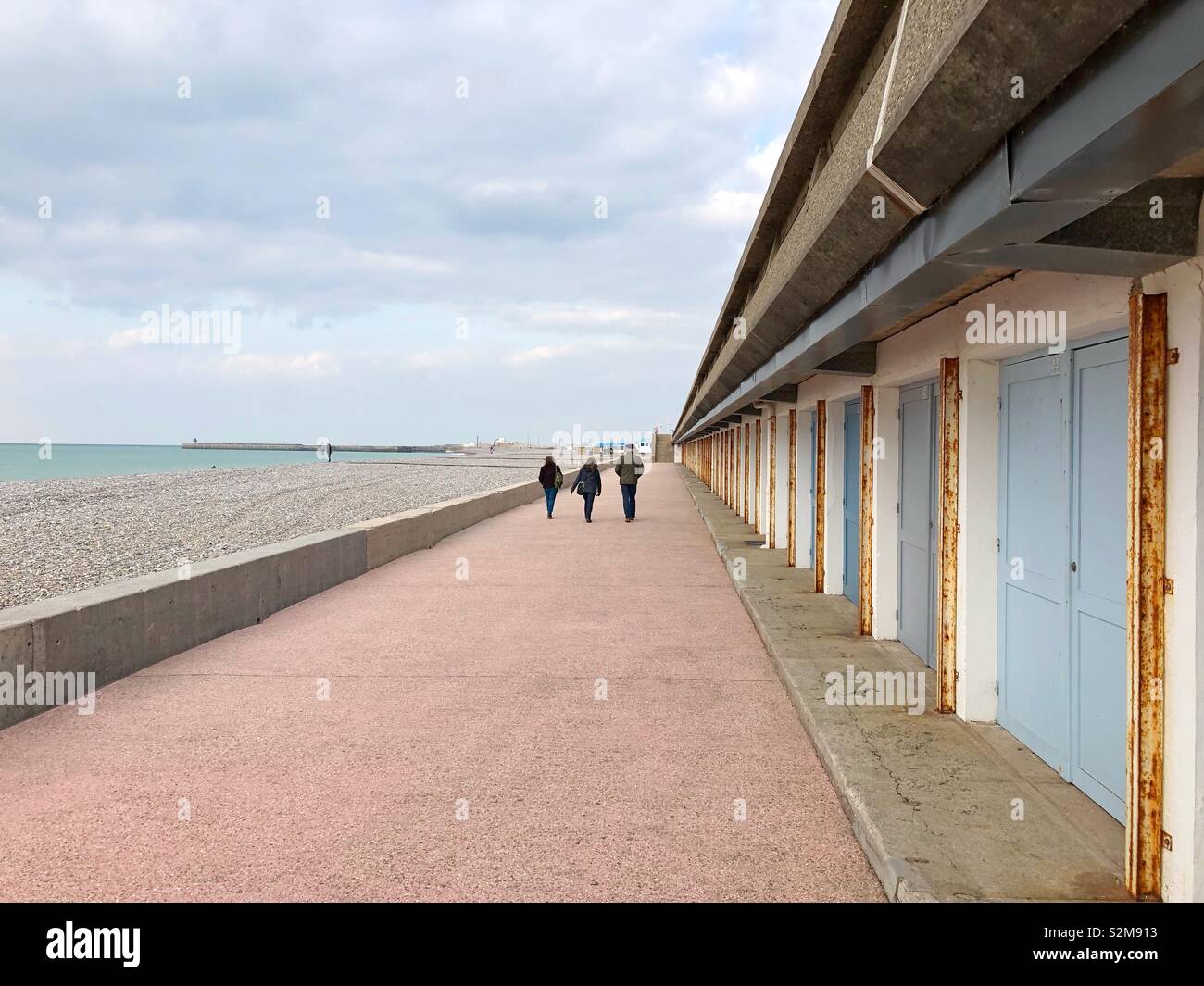 Three persons walking past pastel coloured beach cabins on the Dieppe waterfront off season. - Smartphone Captured Stock Image