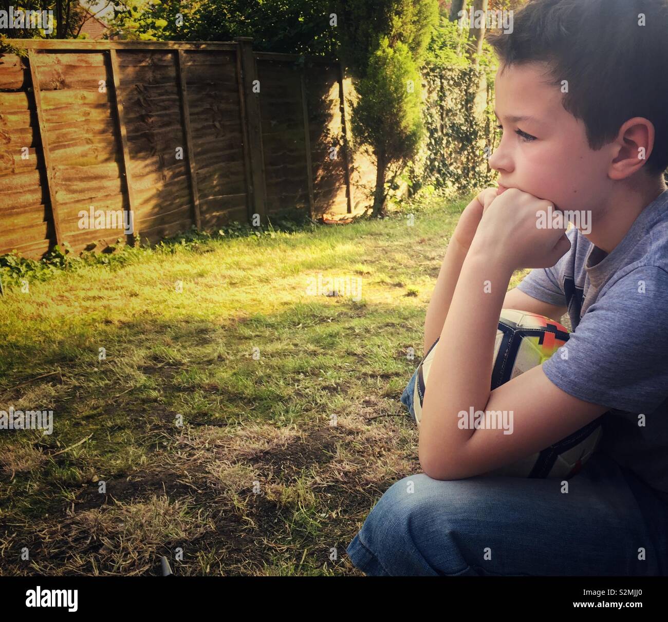A nine-year-old boy sits deep in thought, resting his chin on his hands, while holding a football on his knee. Stock Photo