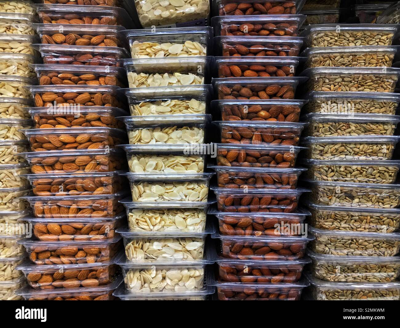Stacked plastic containers full of raw almonds, roasted almonds, sliced almonds, and sunflower seeds on display and for sale at the local produce market. - Smartphone Captured Stock Image