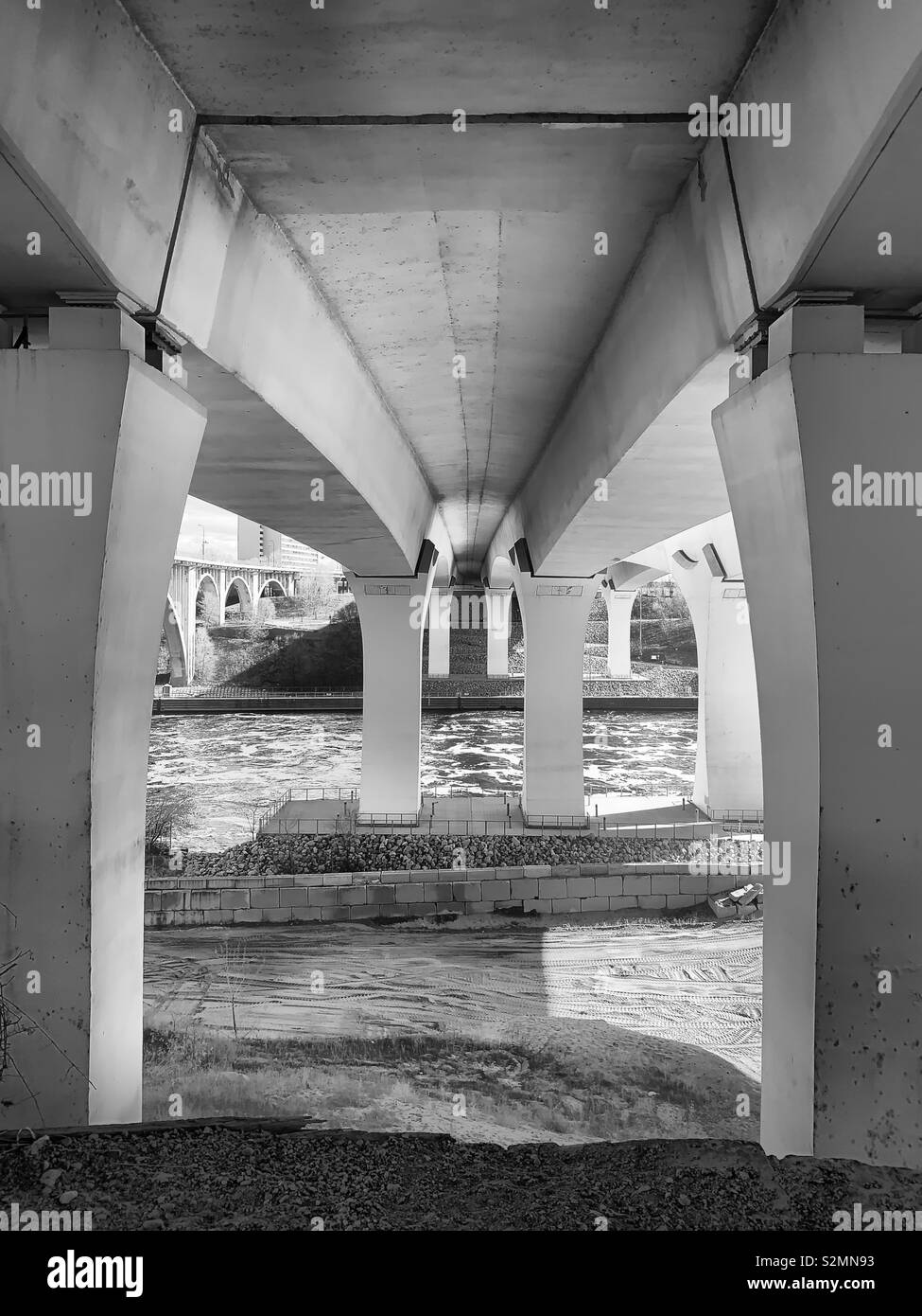 Black and white shot looking up underneath a bridge that spans over a river. Stock Photo