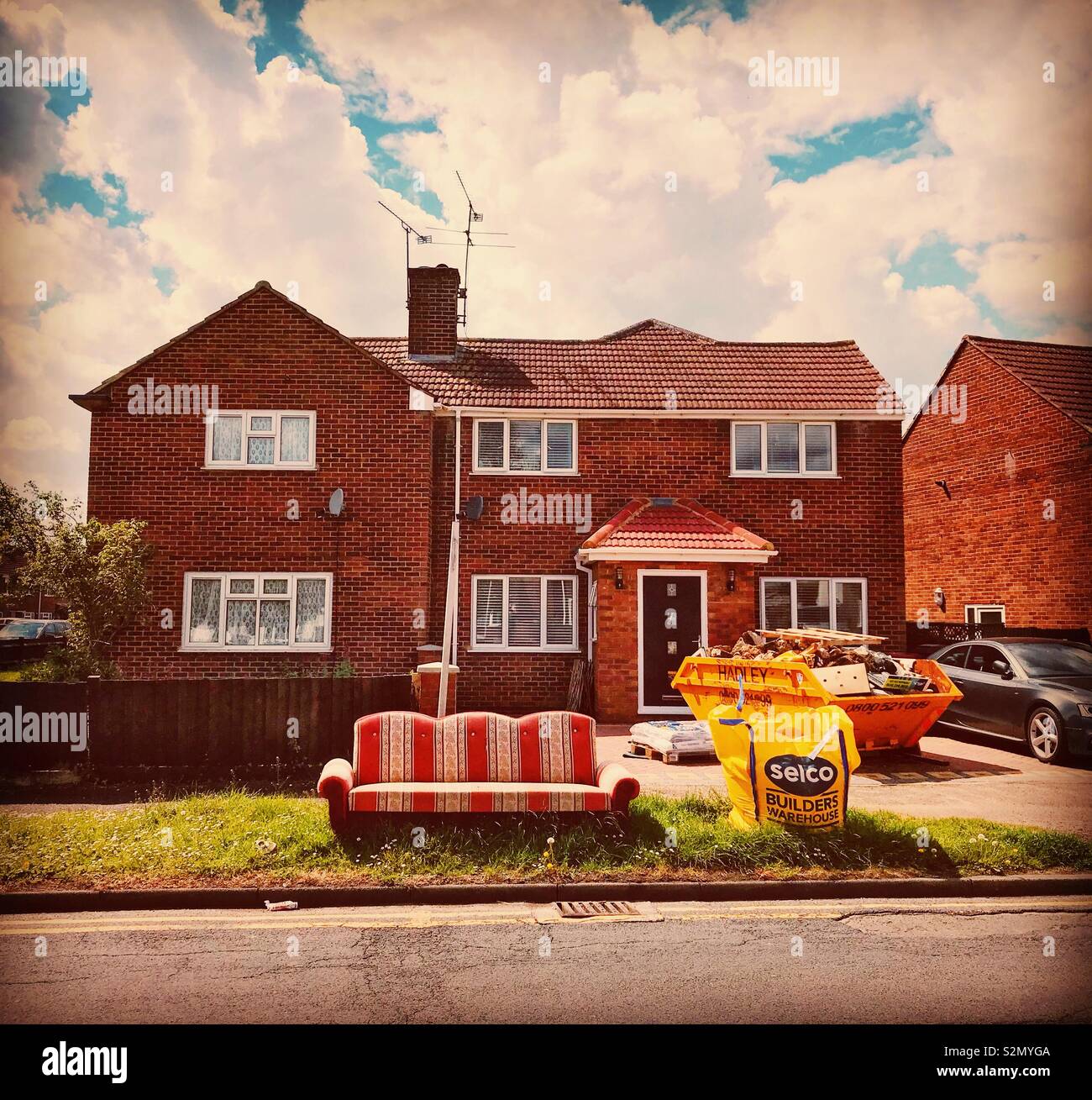 A house undergoing refurbishment with a skip on the drive and an old sofa and bag of building materials on the grass verge. - Smartphone Captured Stock Image