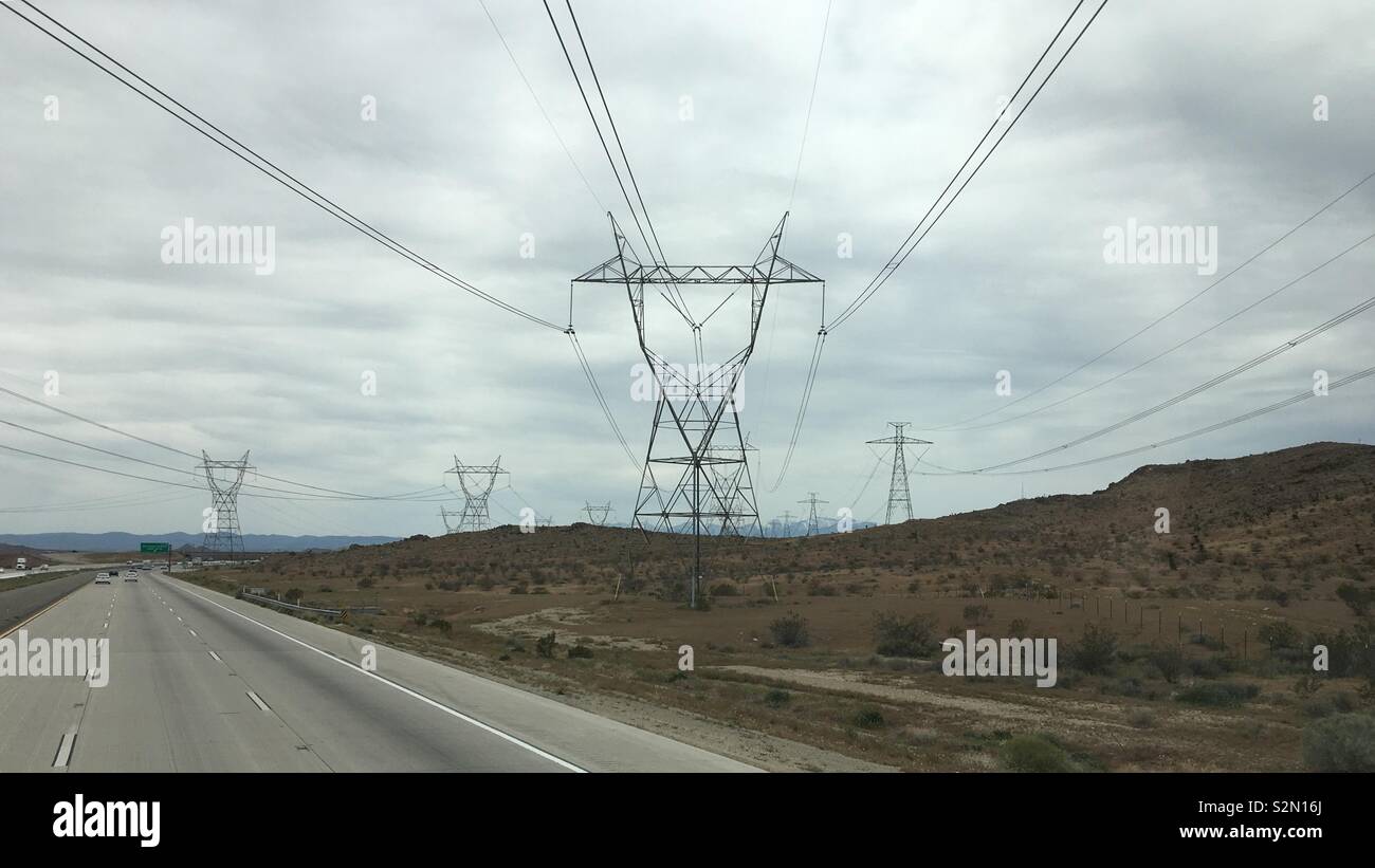 Rows of pylons bearing high tension electric cables crossing freeway and disappearing towards mountains, Apple Valley region, California. Overcast day - Smartphone Captured Stock Image