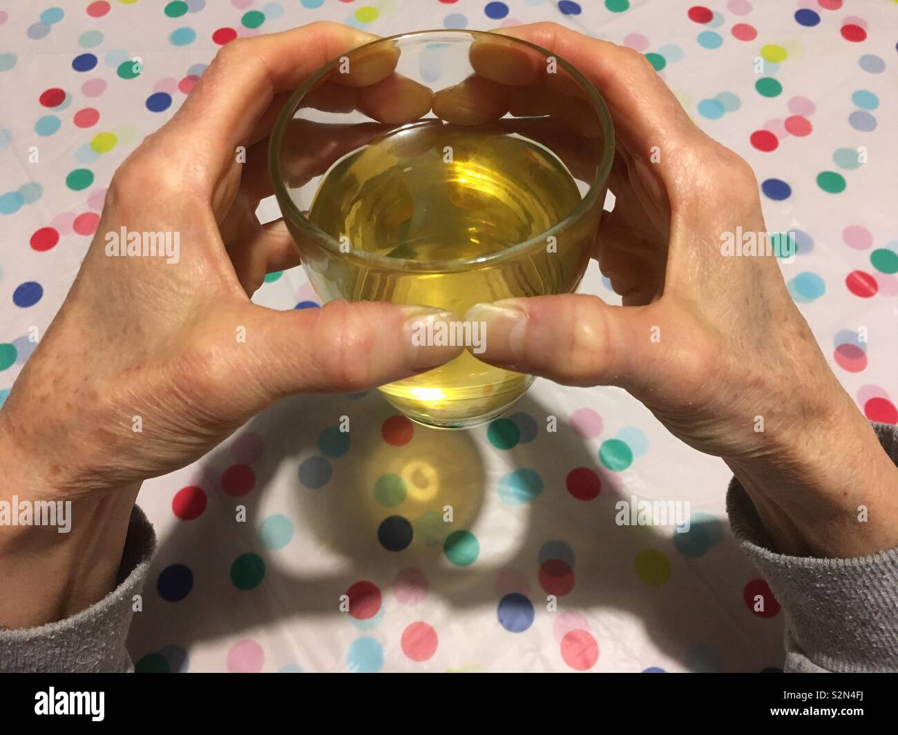 Elderly Caucasian woman’s hands holding a glass full of green tea with lemon myrtle which has been reflected on a plastic pastel polka dotted table cover. Stock Photo