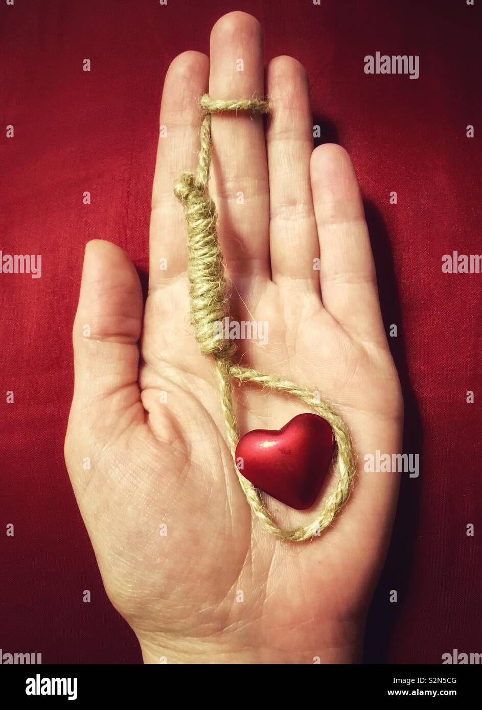 You’re suffocating me: hand holding hangman’s noose around red heart on red background, concept of fatal attraction - Smartphone Captured Stock Image