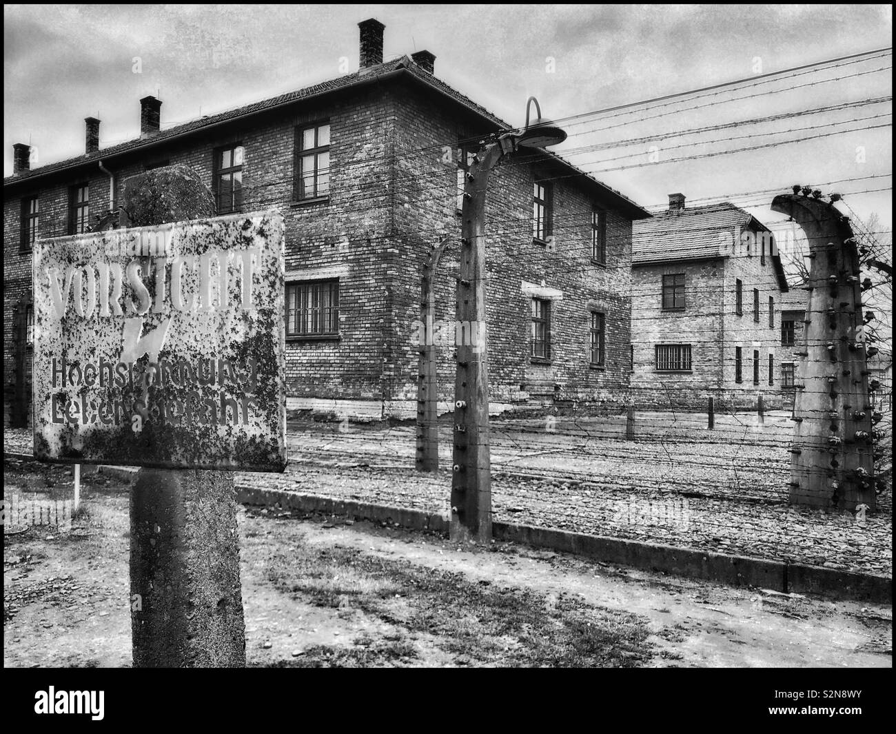 A monochrome image showing some of the buildings & the electric fence at Auschwitz - the infamous Nazi WW2 Concentration Camp in Poland. Thousands died here. Now a UNESCO site & Memorial Museum. © CH. - Smartphone Captured Stock Image