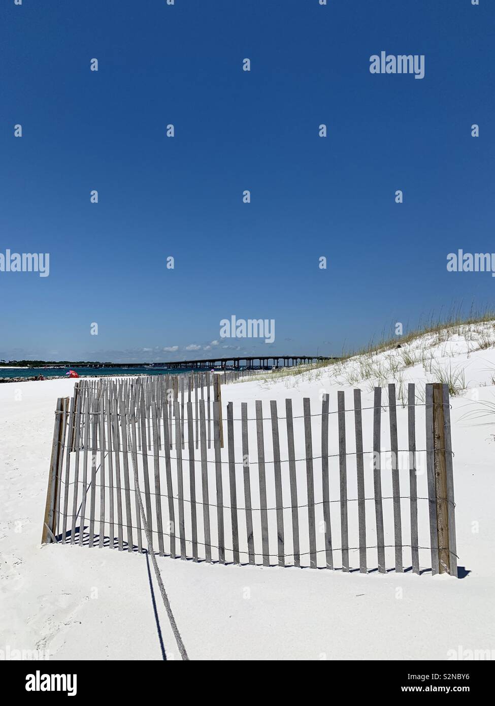 Beach seascape with white sand, fence line, dunes, emerald water and bridge with blue skies - Smartphone Captured Stock Image