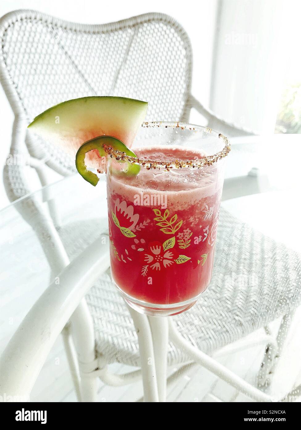 A spicy watermelon margarita in a floral retro glass is surrounded by a white wicker chair for a delicious summer beverage. Stock Photo