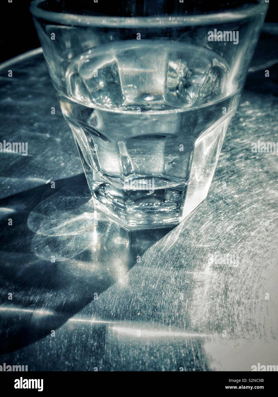 Clear partially full glass of water on metal patio table in bright sunshine in an outdoor urban setting. Stock Photo